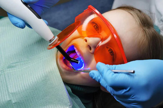 Child in dental clinic wearing orange protective glasses and green bib while dentist uses blue curing light inside mouth.